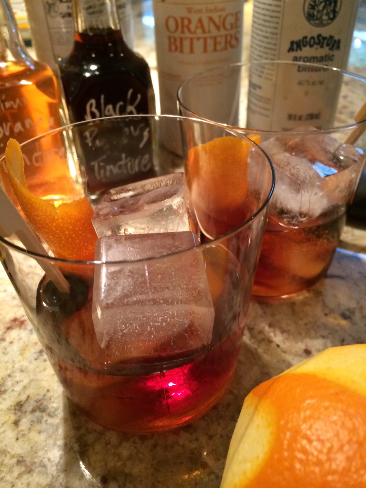 Two cocktails on ice with orange peel, bitters bottles on the bar behind them